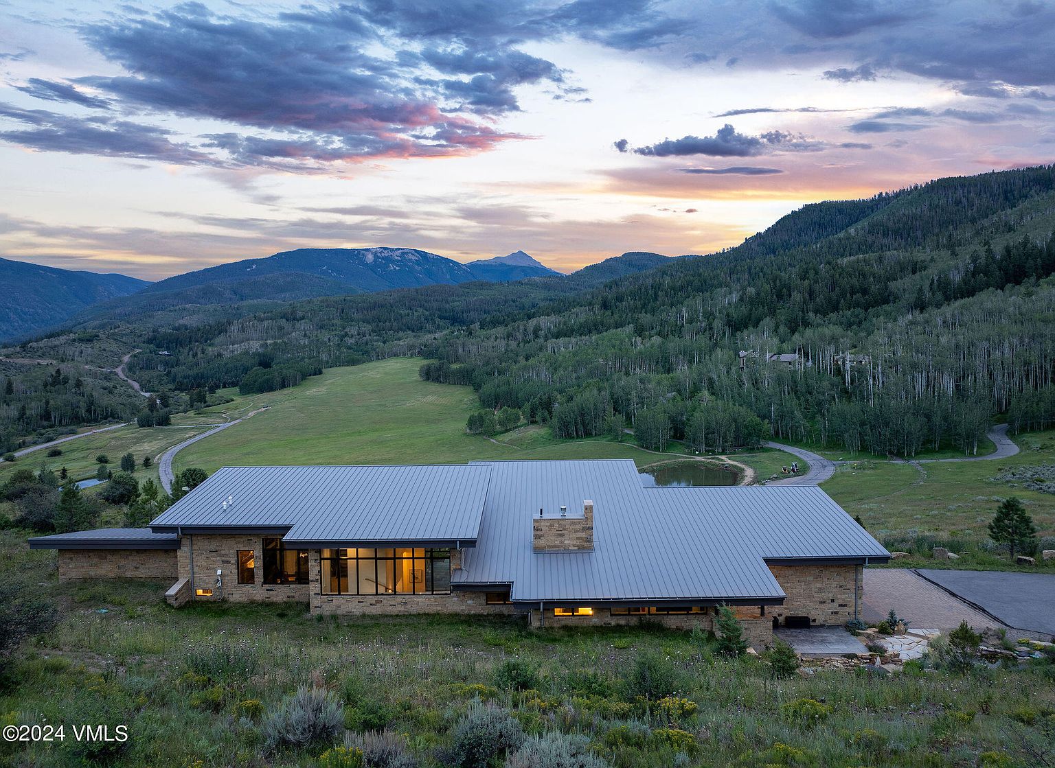 This aerial view showcases a modern, single-story home with a metal roof, nestled in a serene mountain landscape. The property features expansive windows, stone accents, and blends seamlessly with the surrounding greenery and rolling hills. A winding driveway leads to the house, suggesting privacy and seclusion.