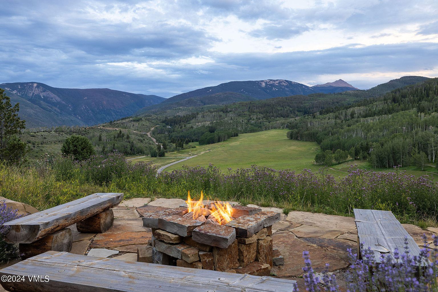 This image showcases a beautifully landscaped outdoor space with a stone fire pit as the focal point, surrounded by wooden benches. The scene offers a serene view of rolling hills and mountains under a cloudy sky, creating an inviting atmosphere for relaxation and outdoor gatherings. The presence of wildflowers adds a touch of natural beauty to the setting.