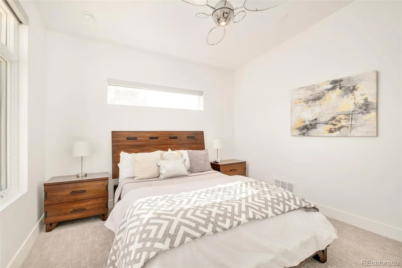 This is a well-lit bedroom featuring a wooden headboard bed with neutral-toned bedding and decorative pillows. Two wooden nightstands flank the bed, each topped with a lamp. An abstract painting hangs on the wall, and a modern ceiling fan is visible above, creating a comfortable and stylish space.