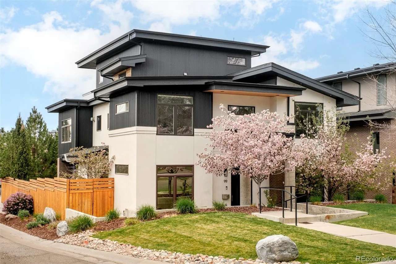 This is a front exterior view of a modern, multi-level home with a striking black and white color scheme. The house features a combination of flat and gabled rooflines, large windows, and a well-manicured lawn with decorative rock landscaping. A flowering tree adds a touch of color and elegance to the property's curb appeal.