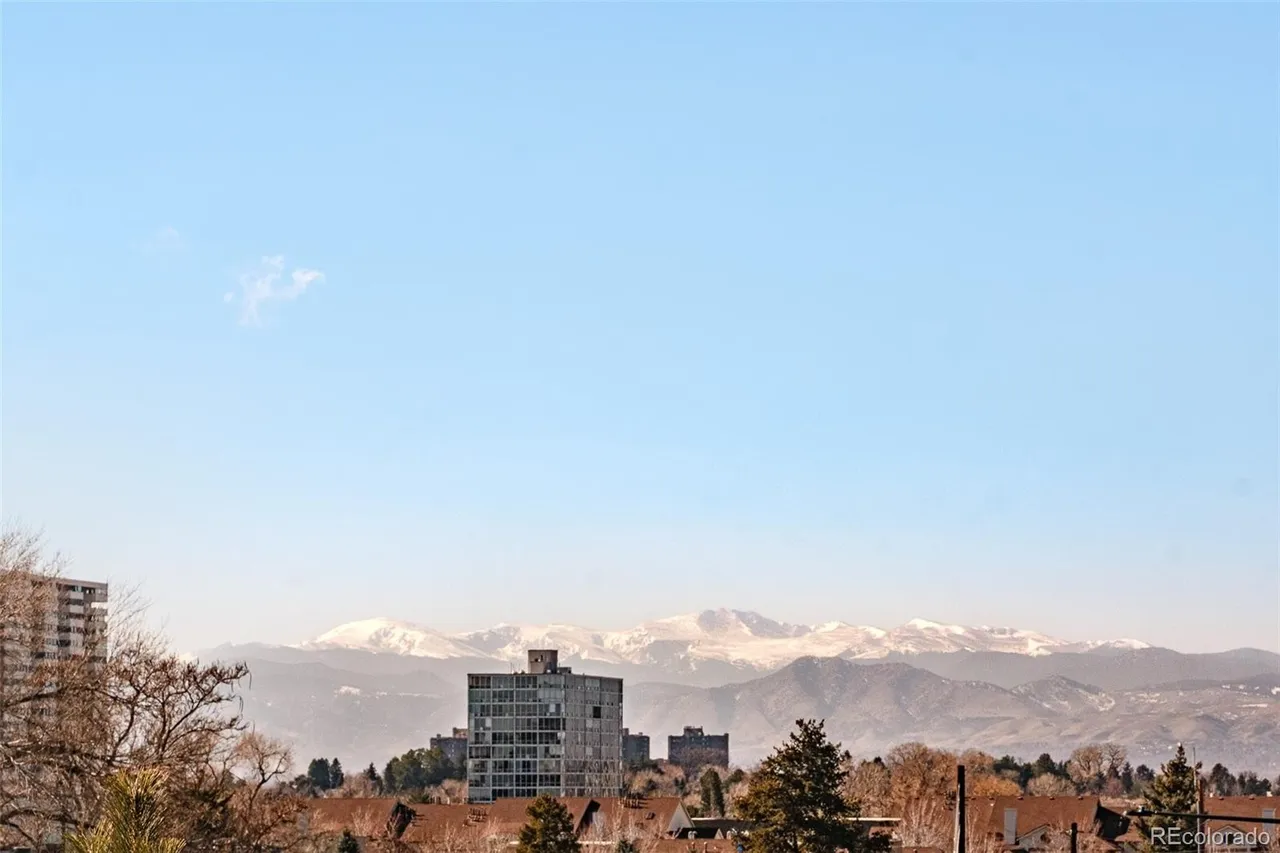 This aerial view showcases a cityscape with a backdrop of snow-capped mountains under a clear blue sky. Several high-rise buildings are visible among lower residential structures and trees, creating a sense of urban living with natural beauty. The image provides a broad perspective, emphasizing the location's proximity to both city amenities and scenic landscapes.