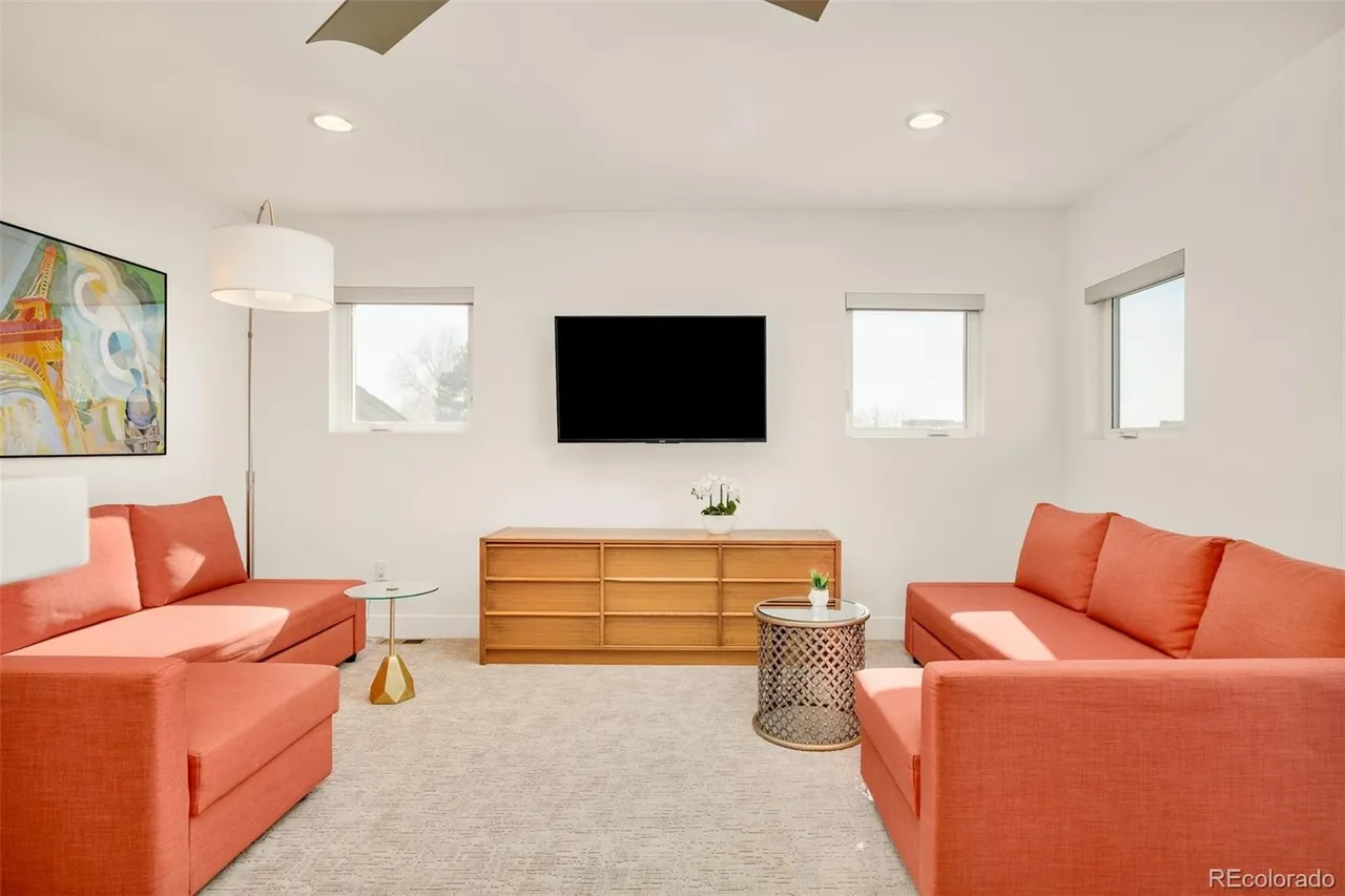 This is an interior shot of a living room featuring two orange sofas facing each other, a wooden media console under a wall-mounted TV, and three windows providing natural light. The room is decorated with modern art and side tables, creating a clean and inviting atmosphere. The perspective is from the center of the room, showcasing the symmetrical arrangement of the furniture.