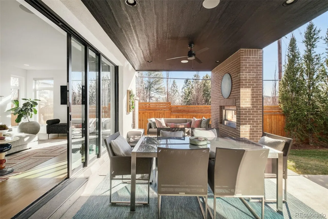 This image showcases a stylish outdoor patio area, featuring a modern dining set with seating for six, set upon a blue patterned rug. The patio is partially covered with a dark wood ceiling and includes a brick fireplace with a circular metal accent. Sliding glass doors lead into the home, offering a seamless transition between indoor and outdoor living spaces.