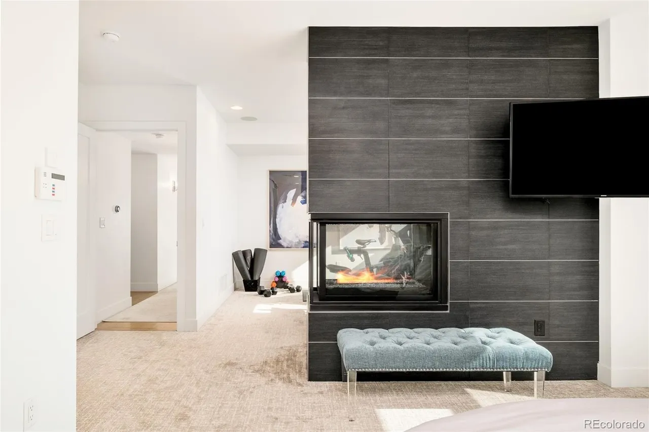 This is a primary bedroom featuring a modern fireplace with a mounted television above. The room has neutral-toned carpet and white walls, leading into a hallway with a glimpse of a home gym area. A tufted bench sits in front of the fireplace, adding a touch of elegance.