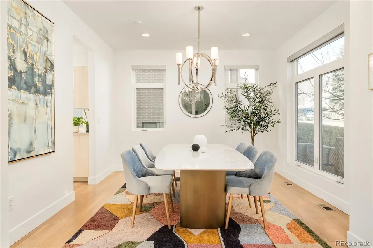 This is a bright and airy dining room featuring a modern aesthetic. A rectangular dining table with a white marble top and a gold base is surrounded by six upholstered chairs in a light blue hue. The room is illuminated by a contemporary chandelier and natural light streaming through the windows, complemented by a colorful geometric rug that adds a touch of vibrancy to the space.