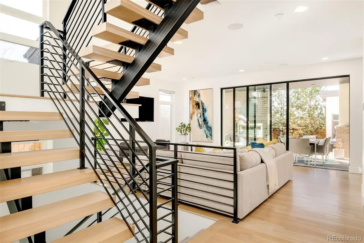 This interior shot showcases a modern living room with a striking staircase featuring wooden steps and a black metal railing. The open floor plan leads to an outdoor patio visible through large sliding glass doors, enhancing the sense of space and light. A neutral-toned sofa and abstract artwork add to the contemporary aesthetic.