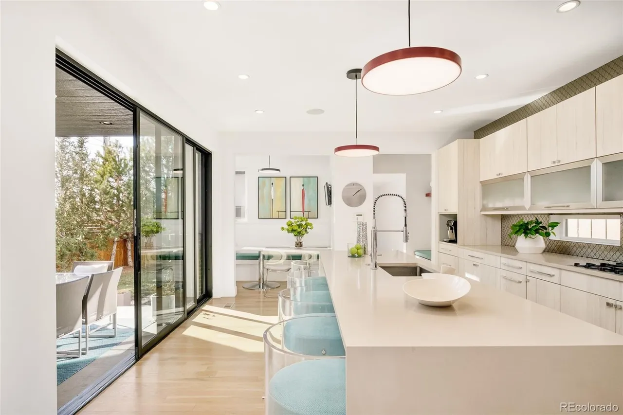 This is a bright, modern kitchen featuring a large island with seating, sleek cabinetry, and stainless steel appliances. The kitchen opens to an outdoor patio through large sliding glass doors, creating a seamless indoor-outdoor living experience. The perspective is from the entrance of the kitchen, showcasing the island and the outdoor view.