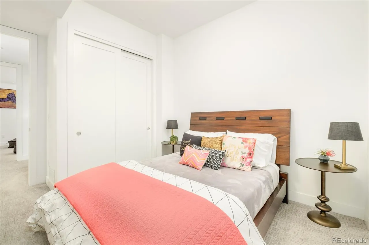 This is a cozy bedroom featuring a wooden headboard, a bed with decorative pillows and a coral blanket, and two side tables with lamps. The room has white walls and a closet with sliding doors, creating a clean and inviting space. The perspective is from the foot of the bed, showcasing the entire room.