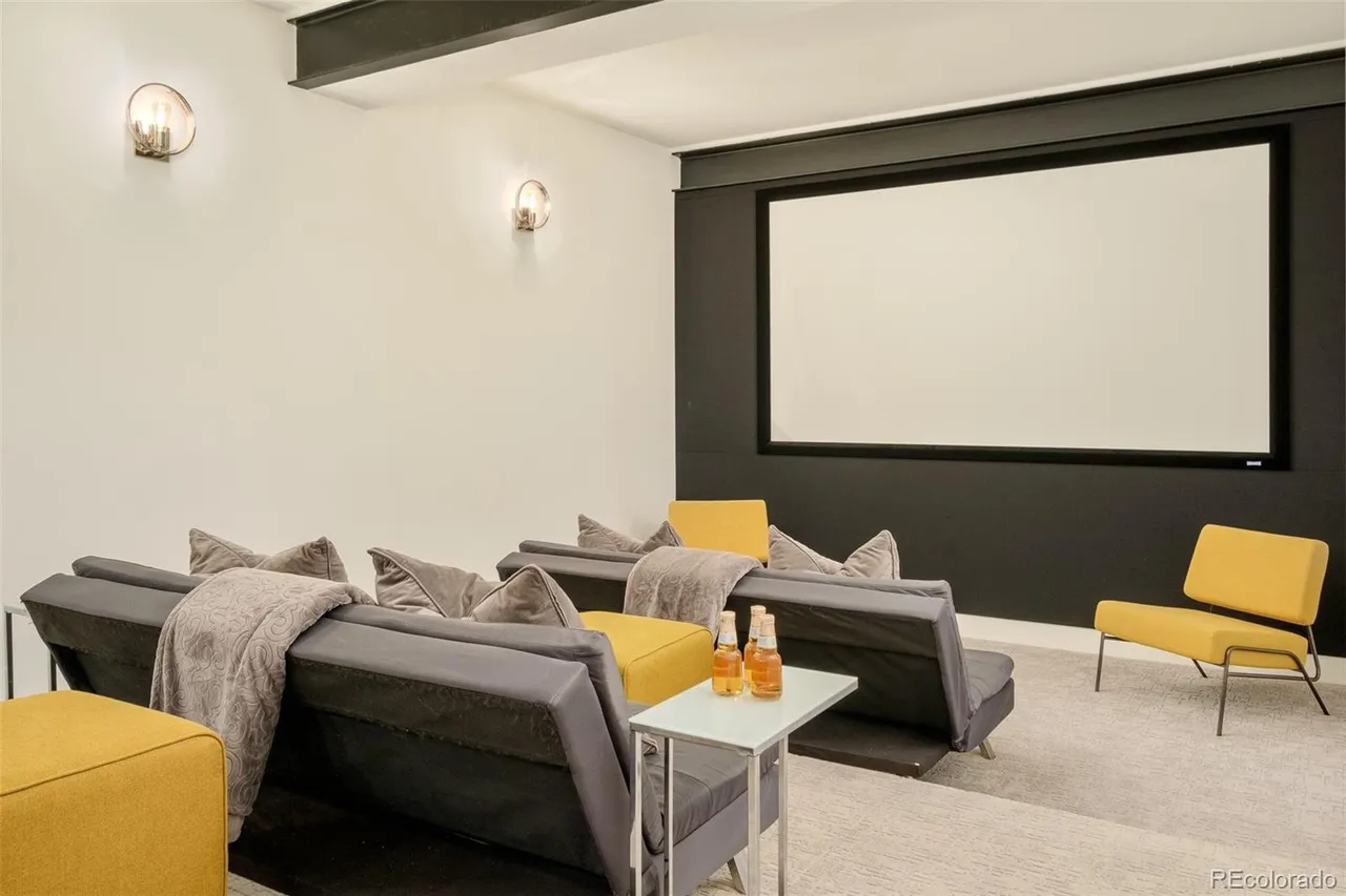 This is an interior shot of a home theater in a basement. The room features tiered seating with gray sofas and yellow accent chairs, facing a large projection screen framed by a dark wall. Wall-mounted sconces provide ambient lighting, and a small side table holds decorative bottles, creating a cozy and inviting entertainment space.