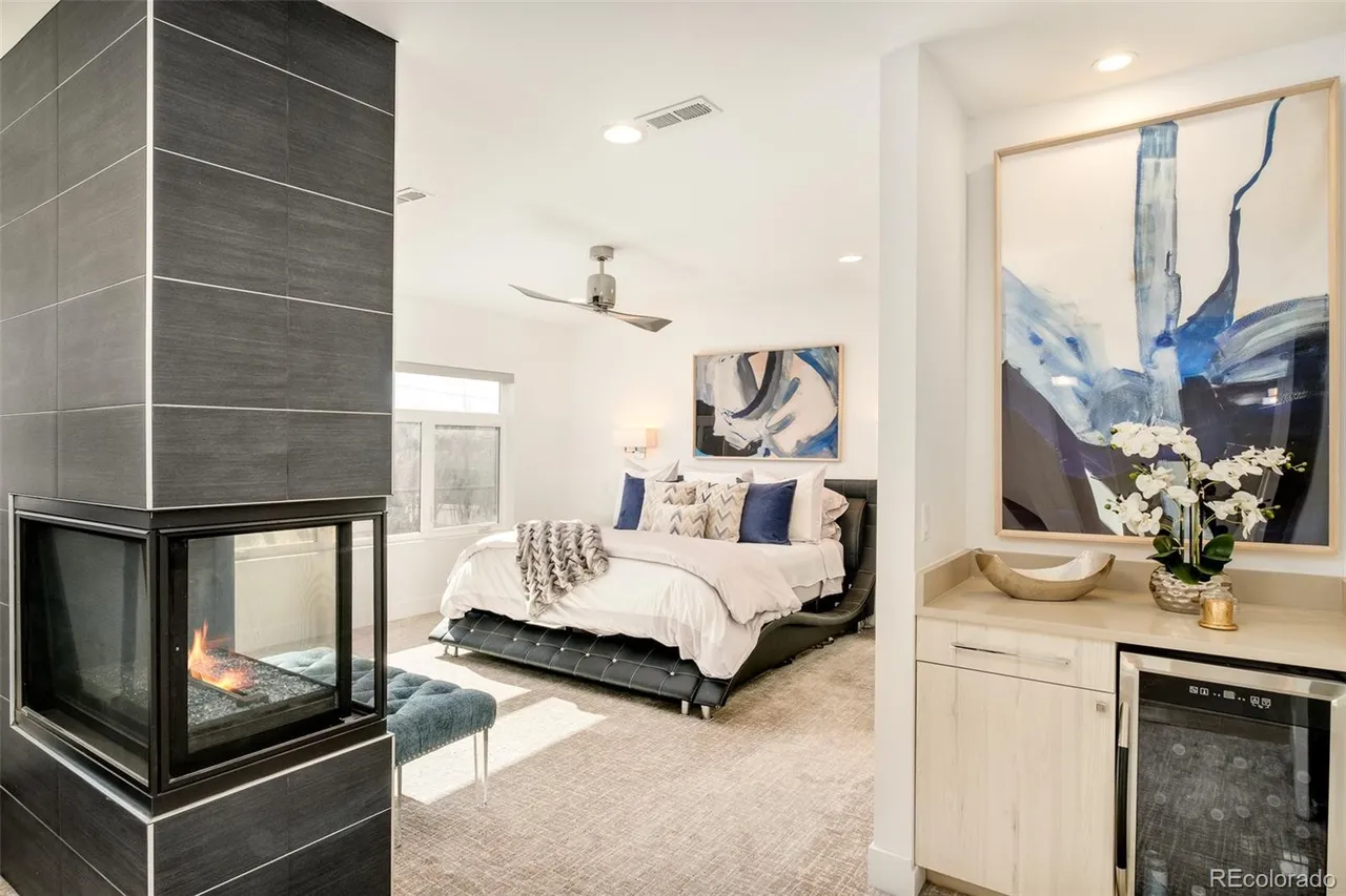 This is a view of a primary bedroom featuring a modern fireplace with dark gray tile, a large bed with white linens and blue accent pillows, and abstract artwork. A built-in bar area with a wine fridge is visible to the right. The room has a contemporary design with a neutral color palette and ample natural light.