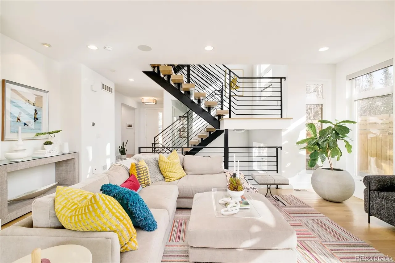 This is a bright and airy living room featuring a large sectional sofa adorned with colorful pillows, a modern staircase with black railings, and a stylish area rug. Natural light floods the space through large windows, highlighting the hardwood floors and creating a welcoming atmosphere. The room is decorated in a contemporary style with clean lines and neutral tones, accented by pops of color.
