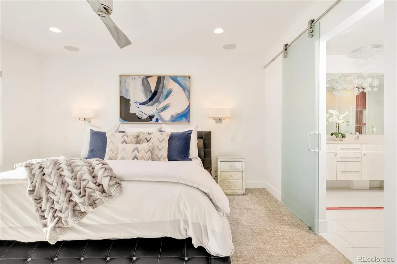 This is a primary bedroom featuring a large bed with white linens, decorative pillows, and a gray throw blanket. An abstract painting hangs above the bed, flanked by sconce lighting. A sliding glass door leads to the primary bathroom, which is visible in the background. The room has a clean, modern aesthetic with white walls and a neutral-toned rug.