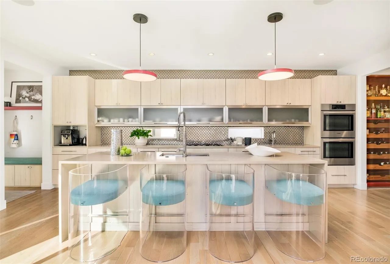 This is a bright and modern kitchen featuring light wood cabinetry, stainless steel appliances, and a large island with a light countertop. The island is surrounded by four clear acrylic bar stools with blue cushions. Two pendant lights with red accents hang above the island, adding a pop of color to the space.