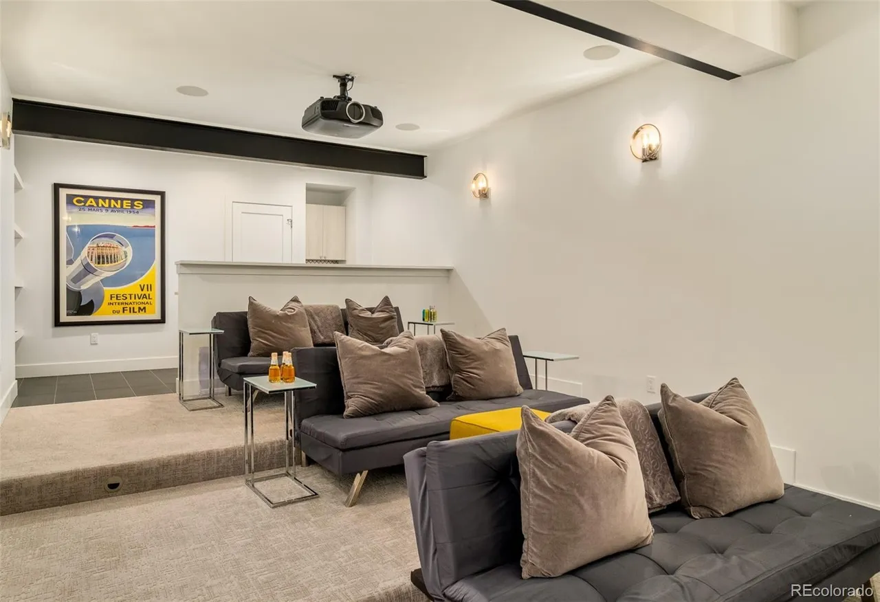 This is an interior shot of a home theater in a basement. The room features tiered seating with gray futons and plush pillows, side tables, and a projector mounted on the ceiling. A framed movie poster adds a touch of cinematic flair, and the overall design creates a cozy and inviting space for entertainment.