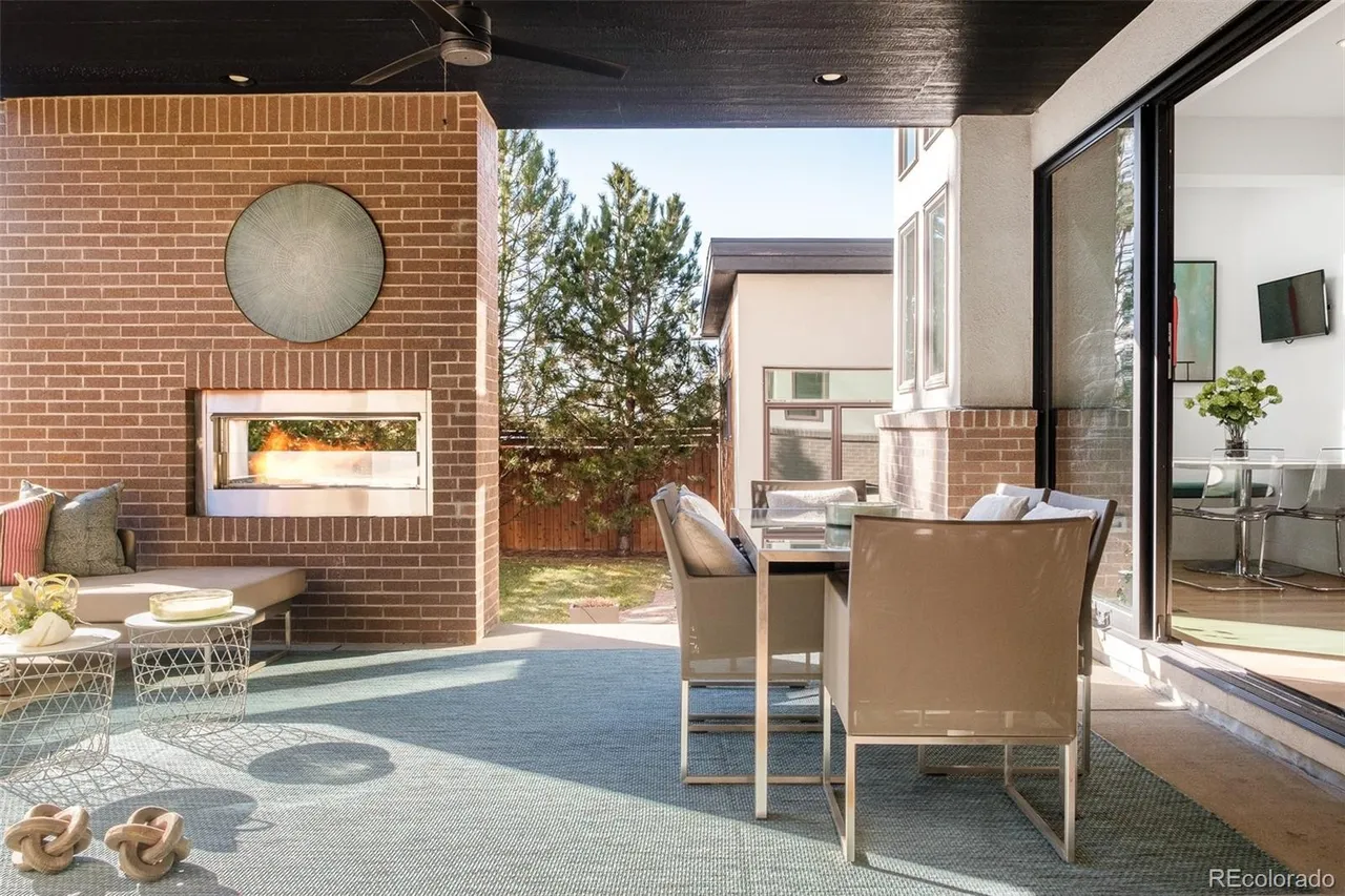 This image showcases a stylish outdoor patio area, featuring a brick fireplace with a decorative metal disc above. The patio is furnished with modern seating and a dining set, all set on a blue-grey outdoor rug. Sliding glass doors lead into the interior, creating a seamless indoor-outdoor living experience.