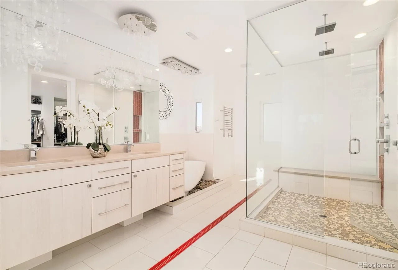 This is a bright and modern primary bathroom featuring a double vanity with light-colored cabinetry and a light countertop. A large mirror spans the length of the vanity, reflecting a decorative chandelier and a glimpse into a walk-in closet. The bathroom also includes a glass-enclosed shower with a built-in bench and a unique pebble-filled area near the bathtub, adding a spa-like touch.