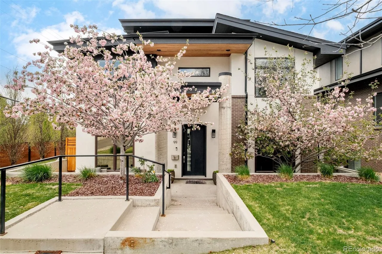 This is a front exterior view of a modern two-story home with a striking architectural design. The facade features a combination of stucco and brick elements, complemented by a dark-colored roof and window frames. Landscaping includes flowering trees and well-maintained lawn, enhancing the curb appeal and creating a welcoming atmosphere.