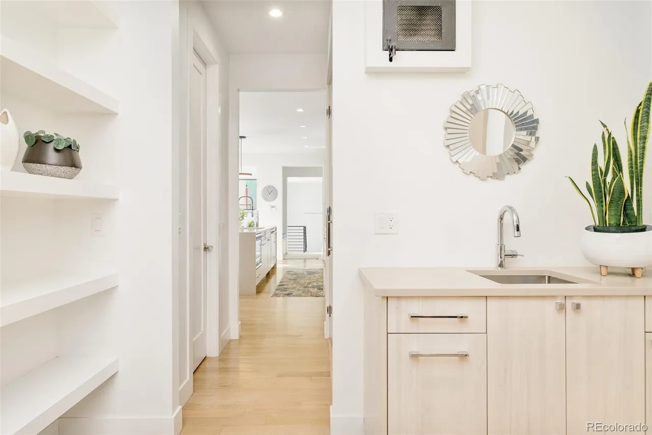 This interior shot showcases a bright hallway with light wood flooring, leading into a kitchen area. The hallway features built-in shelving on the left, a doorway, and a small wet bar area with a sink, mirror, and cabinetry on the right. The overall impression is clean, modern, and well-lit.