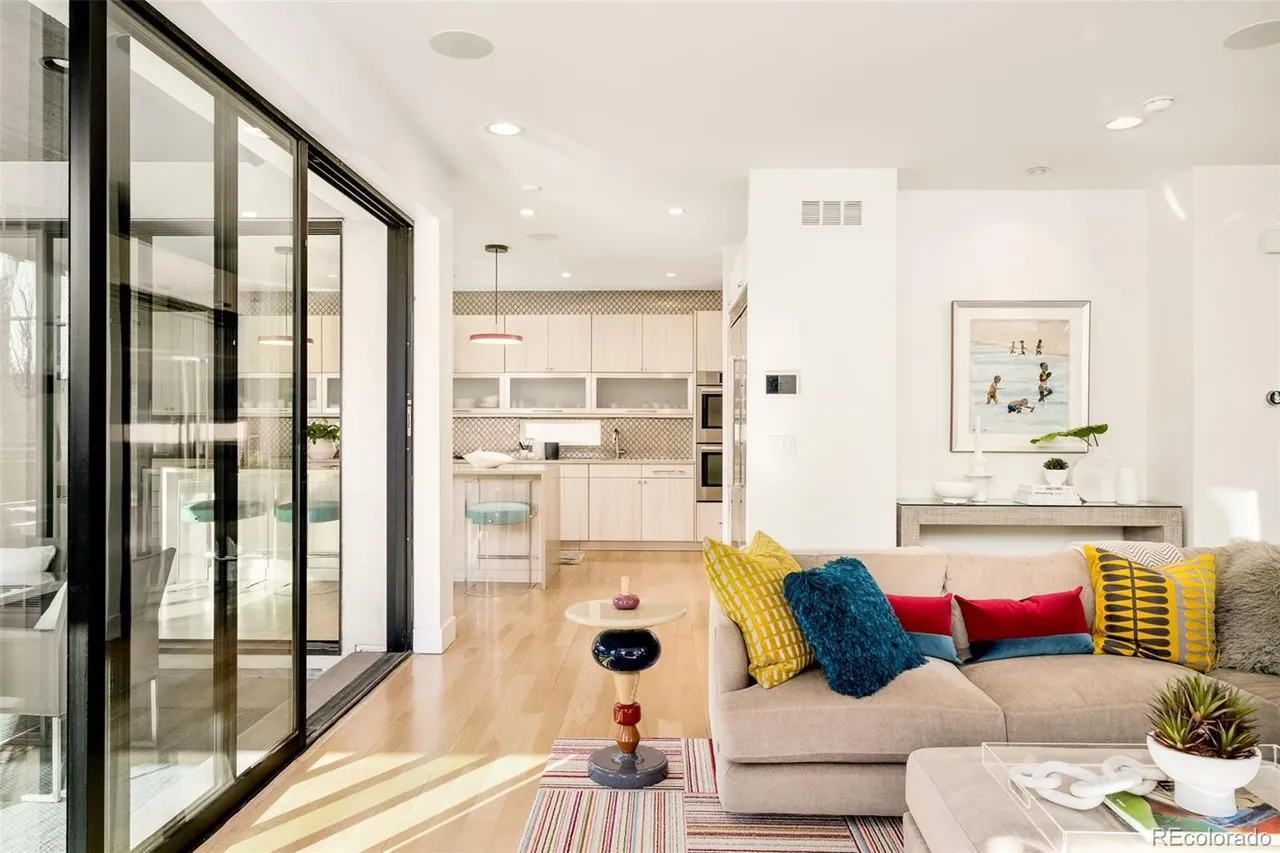 This is an interior shot of a modern living room that seamlessly transitions into a kitchen area. The living room features a comfortable sectional sofa adorned with colorful pillows, a stylish side table, and a striped rug. Large sliding glass doors provide ample natural light and a view to the outside, while the kitchen in the background showcases light wood cabinetry and a breakfast bar.