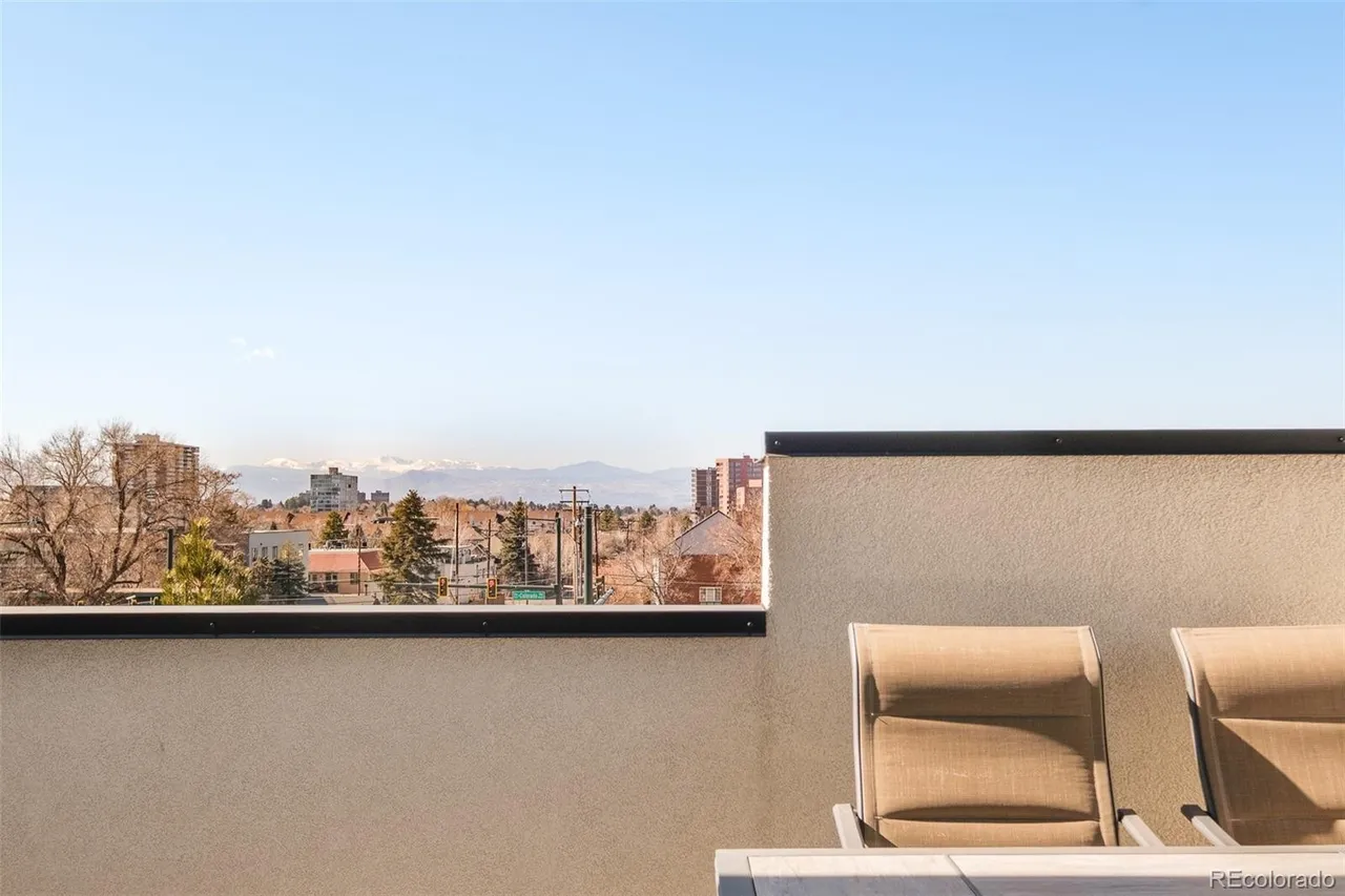 This image showcases a modern patio or balcony with a stunning city and mountain view. The space features a low wall with a dark trim, providing a sense of privacy without obstructing the scenery. Two lounge chairs are partially visible, suggesting a relaxing outdoor seating area perfect for enjoying the vista.