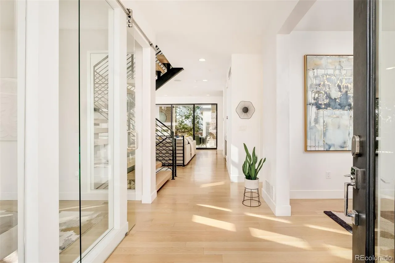 This interior shot showcases a bright and modern hallway with light hardwood flooring and white walls. A glass-enclosed staircase is visible on the left, while an abstract painting and a potted plant add visual interest to the right. The hallway leads to a living area with a sliding glass door, creating an open and airy feel.