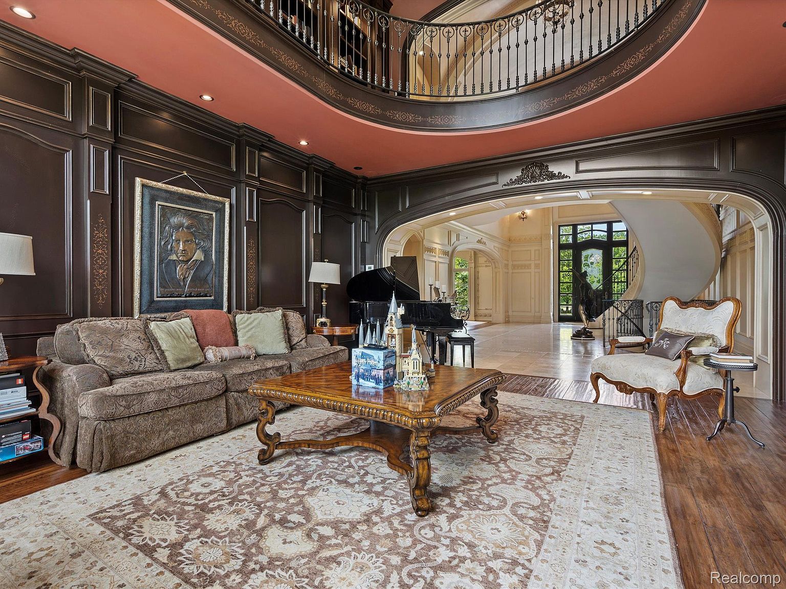 This grand living room features dark wood paneling, a salmon-colored ceiling, and a wrought-iron balcony overlooking the space. A large area rug anchors the room, complementing the ornate coffee table and comfortable sofa. The room opens to a grand foyer with a spiral staircase and a glimpse of the exterior through the front door, creating a luxurious and inviting atmosphere.