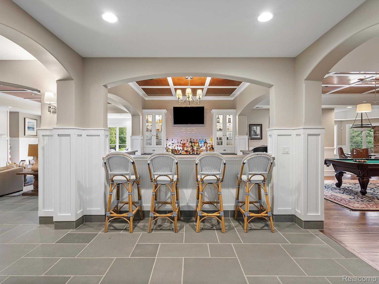 This interior shot showcases a stylish basement bar area, complete with four bar stools, a well-stocked bar, and a television. The space is defined by elegant archways, wainscoting, and a coffered ceiling, creating a sophisticated and inviting atmosphere. The flooring is a large format tile, and the adjacent room features a pool table.