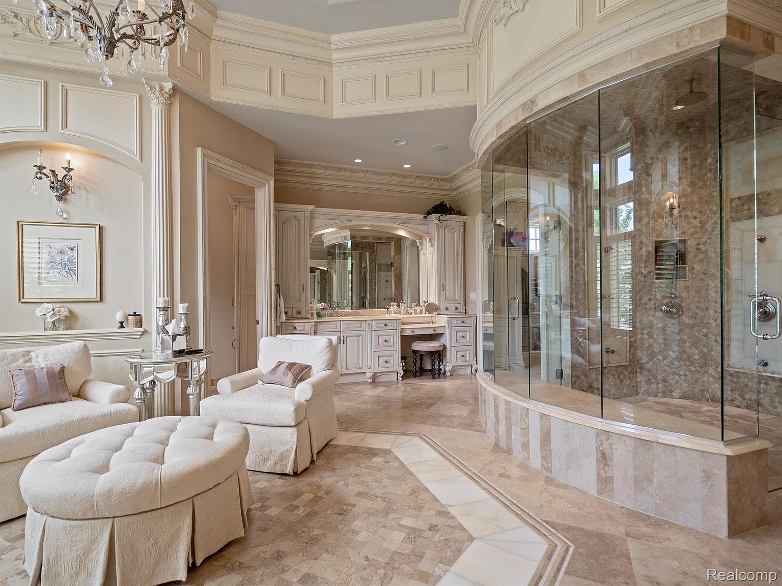 This is a luxurious primary bathroom featuring a large, curved glass shower enclosure with mosaic tile walls and multiple shower heads. The bathroom includes a vanity area with custom cabinetry, a makeup table, and a seating area with a plush sofa and ottoman. The flooring is a patterned tile, and the overall design exudes elegance and sophistication.