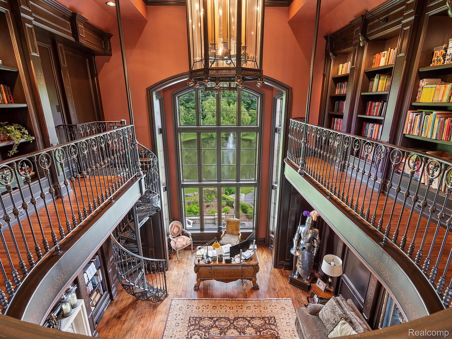 This is a stunning interior shot of a grand home office or study, featuring a two-story space with a large window overlooking a lake. The room is lined with bookshelves on both sides, accessible by a spiral staircase and a balcony. A large desk sits in the center of the room, and a suit of armor stands guard, adding a touch of old-world charm.