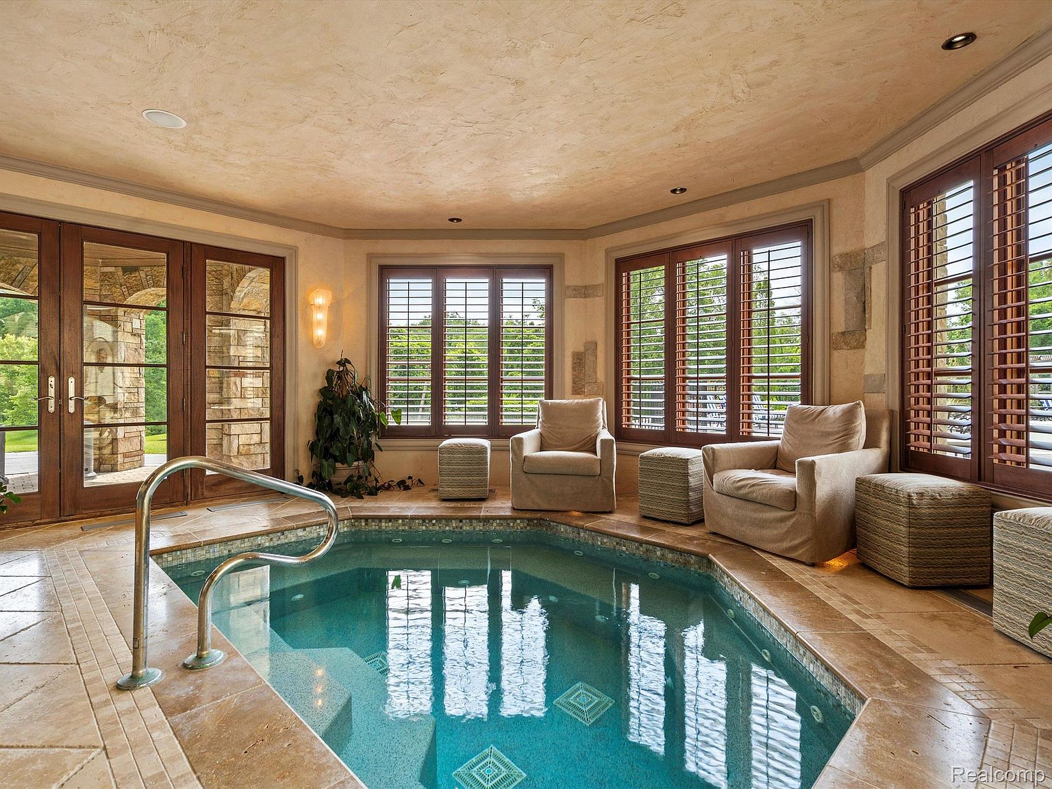 This interior shot showcases a luxurious indoor spa area featuring a tiled hot tub with a stainless steel handrail. The space is well-lit by natural light streaming through multiple windows with wooden shutters, complemented by elegant wall sconces. Comfortable armchairs and ottomans are arranged around the spa, creating a relaxing and inviting atmosphere.