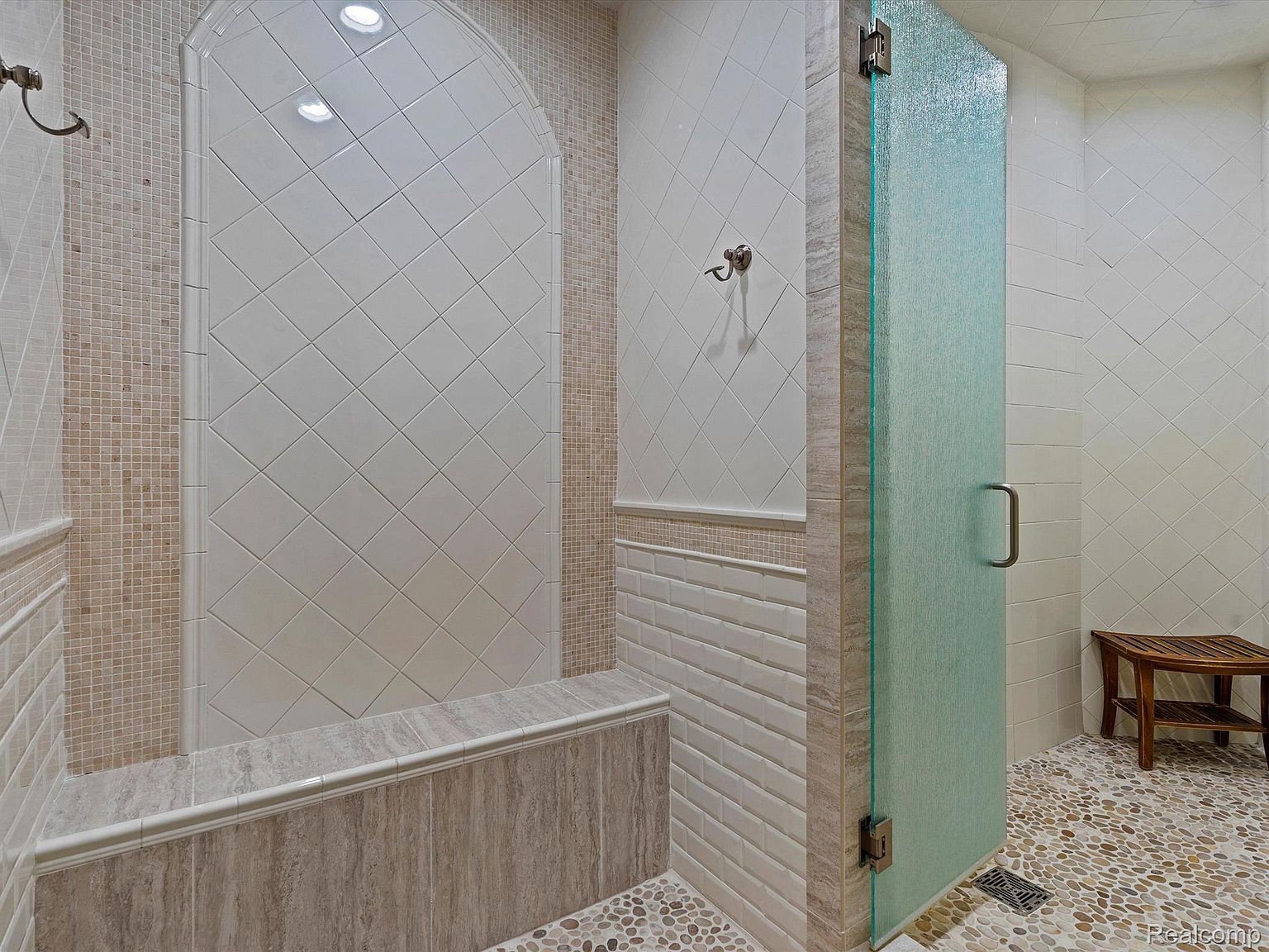 This is a well-appointed primary bathroom featuring a walk-in shower with a glass door and tiled walls. The shower area includes a built-in bench and decorative tile accents. The floor is covered in pebble-style tiles, and a wooden stool sits near the shower entrance, adding a touch of warmth to the space.