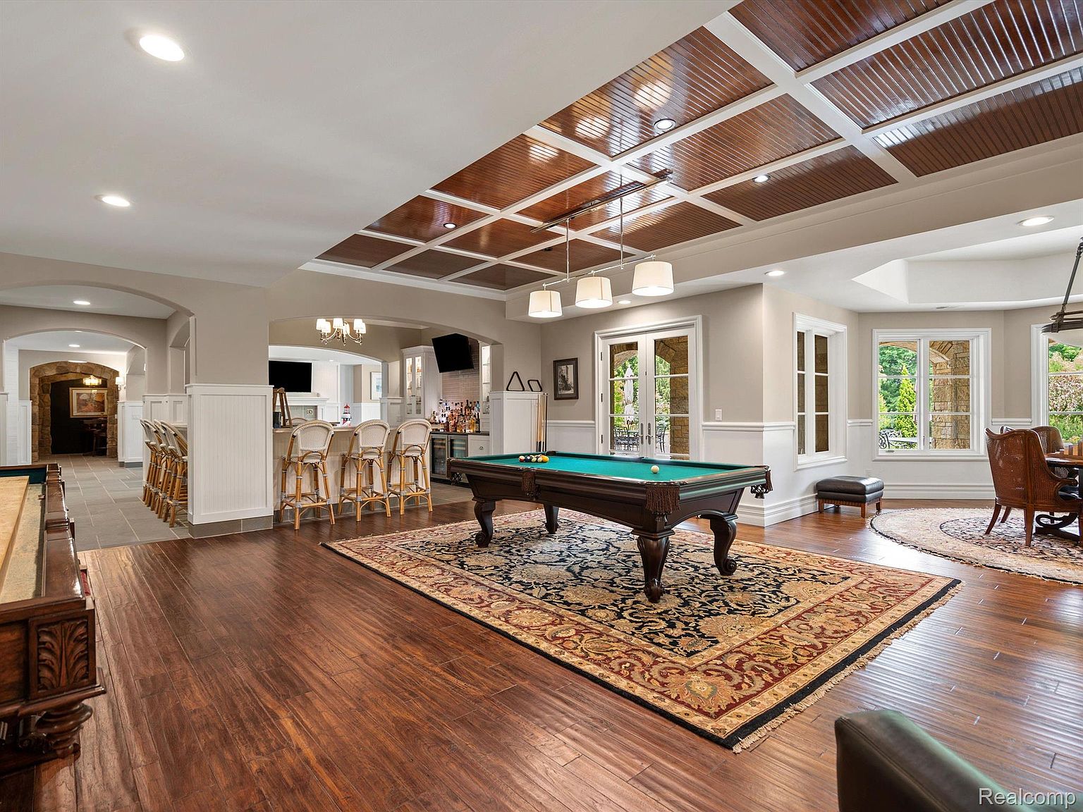 This is a spacious basement featuring a pool table as a focal point, set upon an ornate rug. The room includes a bar area with seating, a dining area with large windows, and a coffered ceiling with recessed lighting. The hardwood flooring and neutral wall colors create a warm and inviting atmosphere, perfect for entertaining.