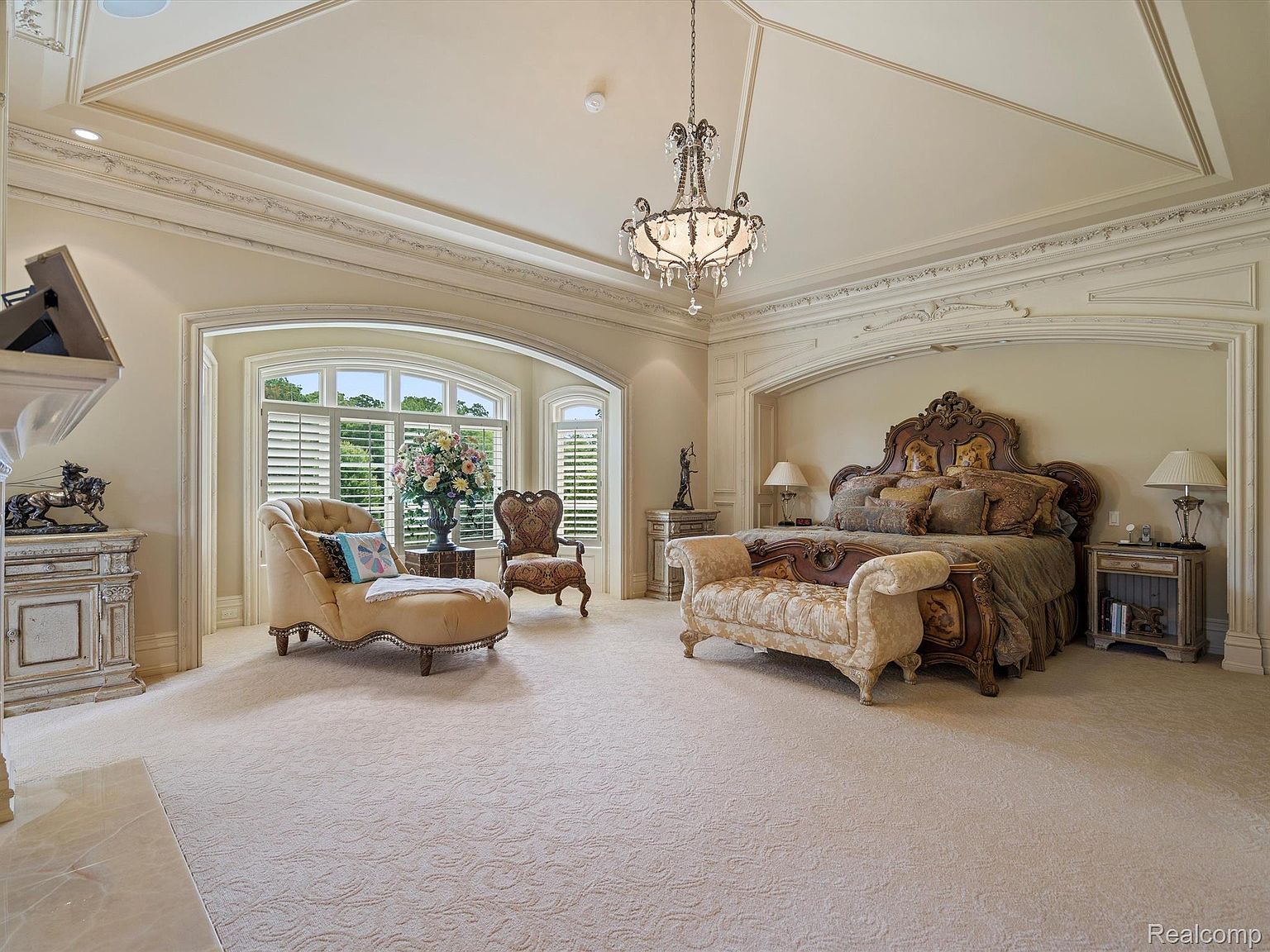 This is a luxurious primary bedroom featuring a large ornate bed with a decorative headboard and matching bench. The room is spacious with a high, detailed ceiling, a crystal chandelier, and an arched window area with a chaise lounge. The overall impression is one of opulence and comfort, perfect for a high-end real estate listing.