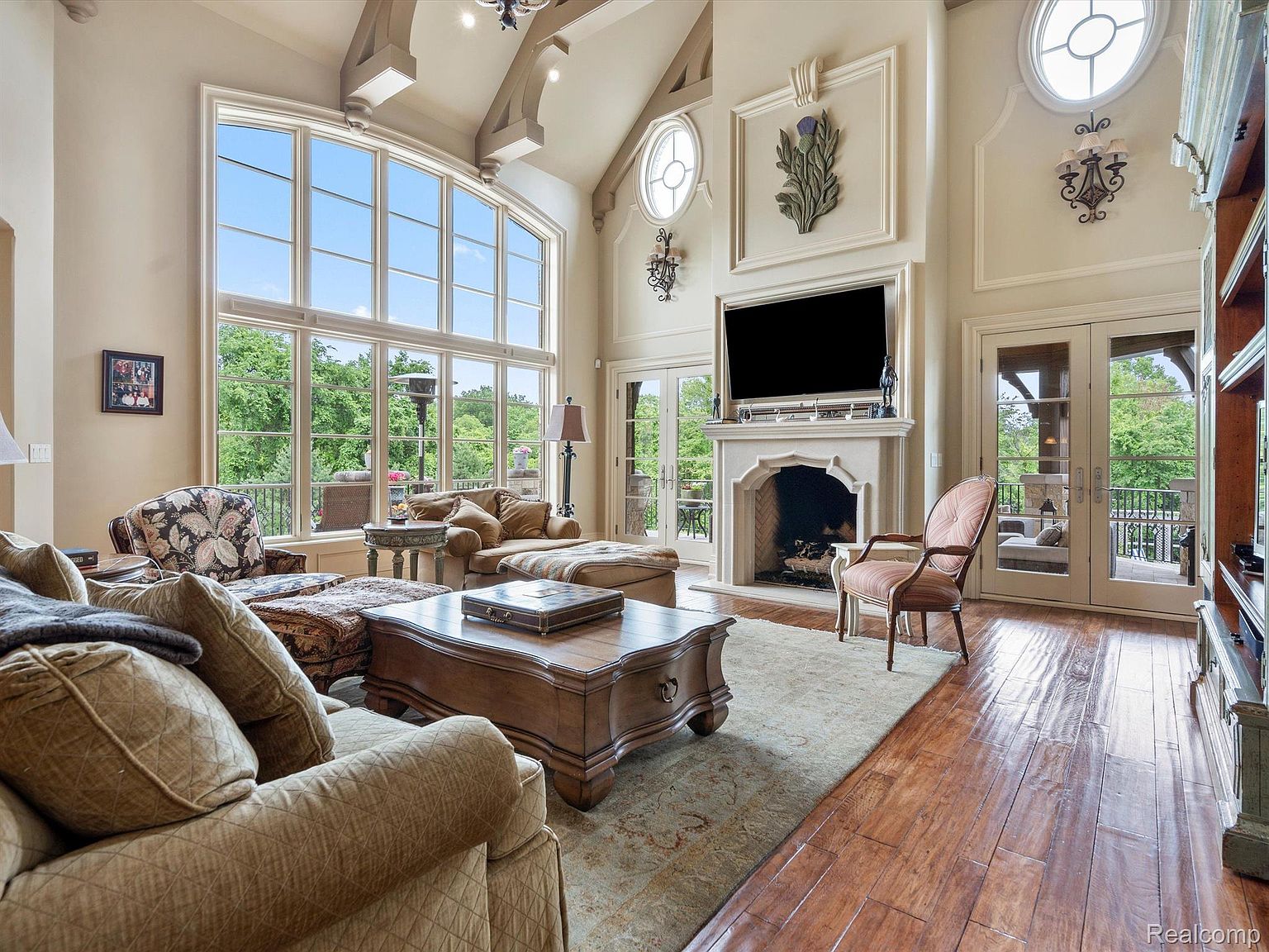 This is a spacious living room featuring a large window with a view of the outdoors. The room is furnished with comfortable seating, a fireplace with a television above it, and hardwood floors. The high ceilings and architectural details add to the luxurious feel of the space.
