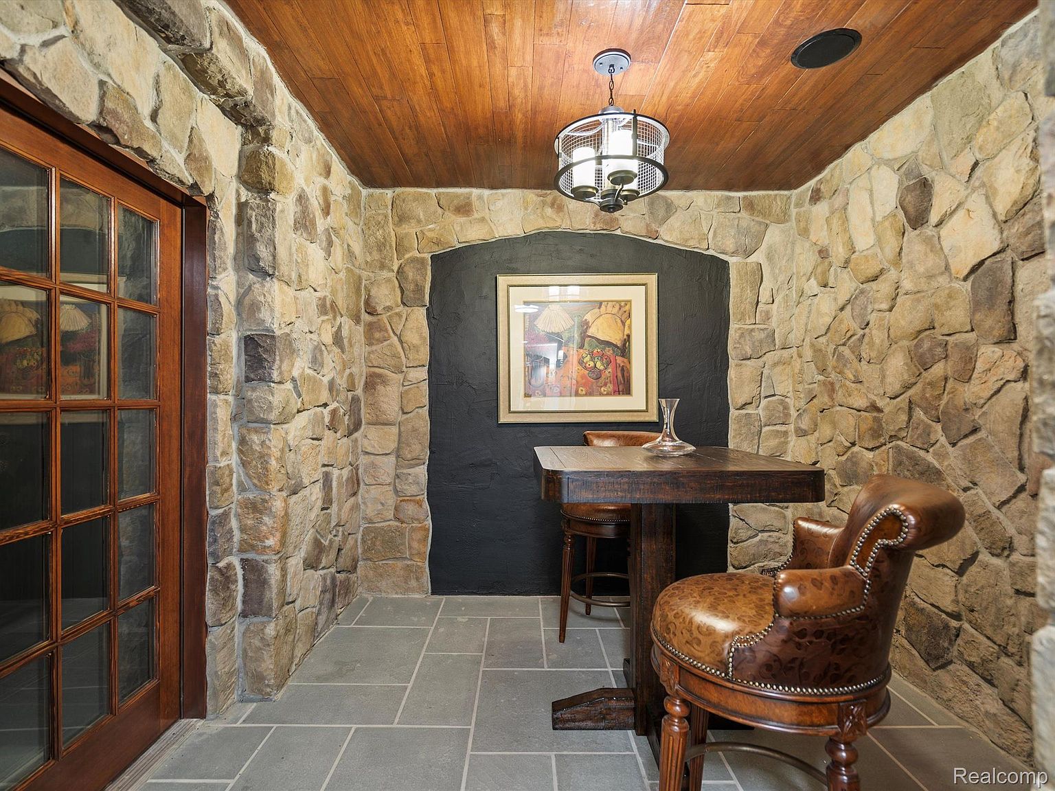This is an interior shot of a rustic wine cellar featuring stone walls and a wooden ceiling. A wooden table with a decanter and chairs are set against a dark accent wall, illuminated by a decorative chandelier. The space exudes a cozy and sophisticated ambiance, perfect for wine tasting and storage.