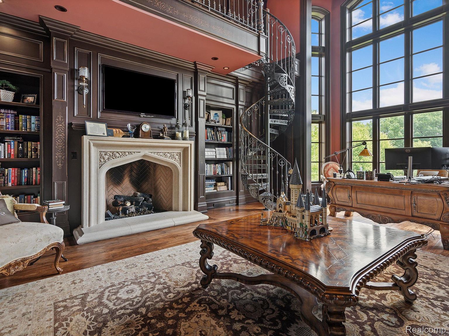 This is a luxurious home office featuring dark wood paneling, built-in bookshelves, and a grand stone fireplace with a mounted television above. A spiral staircase leads to an upper level, and a large window provides natural light. The room is furnished with an ornate desk and a large, decorative coffee table, creating an atmosphere of sophistication and productivity.