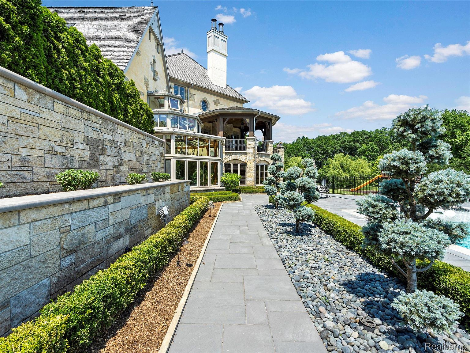 This image showcases the rear exterior of a luxurious home, emphasizing its meticulously landscaped yard and architectural details. A stone walkway leads towards the house, flanked by manicured hedges, decorative rocks, and sculpted trees. The house features a combination of stone and stucco, with a glass-enclosed sunroom and a covered balcony, suggesting a blend of modern and traditional design elements.