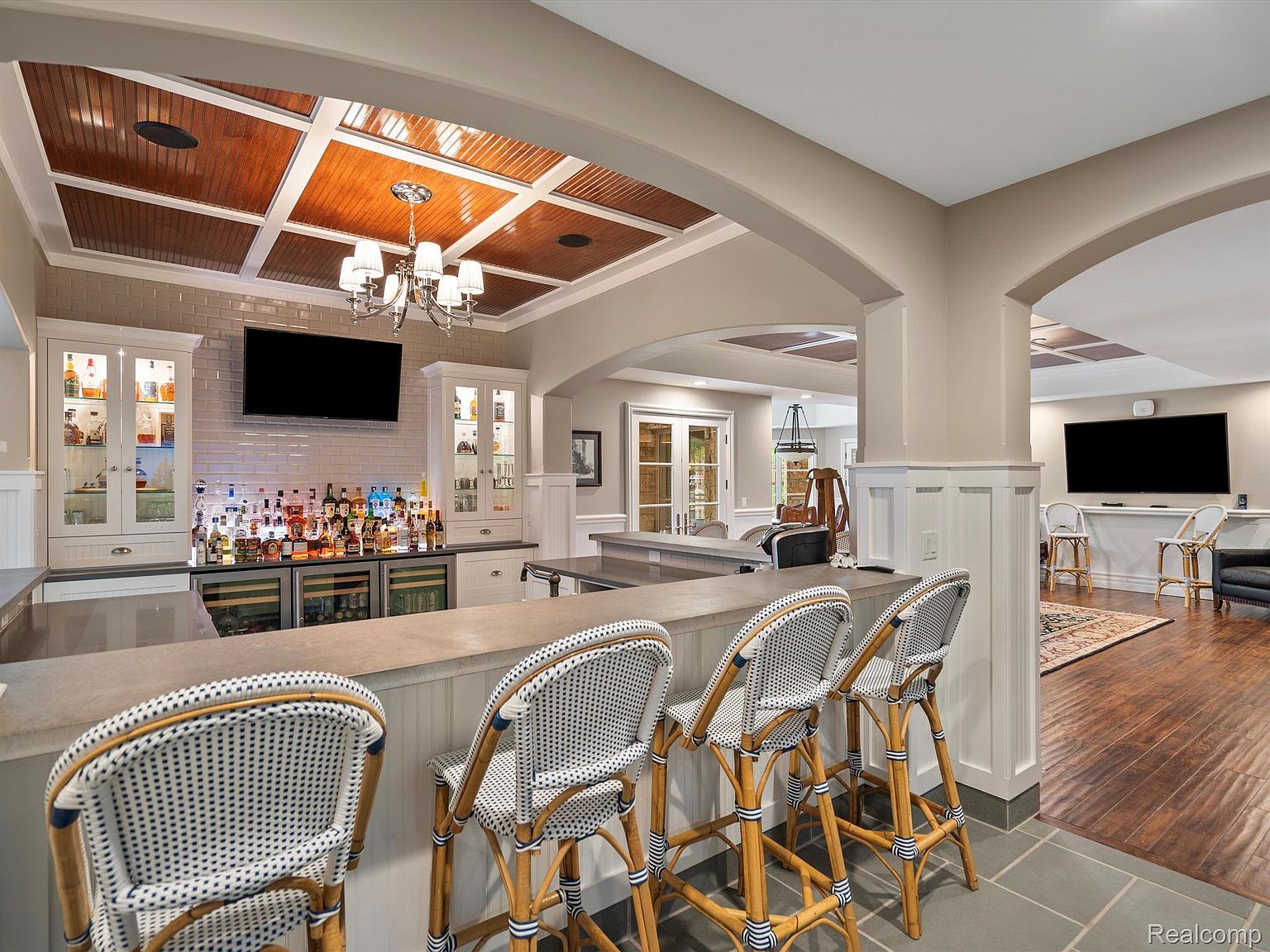 This interior shot showcases a stylish basement bar area, complete with a well-stocked bar, wine coolers, and comfortable seating. The coffered ceiling with wood panels adds a touch of elegance, while the adjacent lounge area with a TV provides a relaxing space for entertainment. The overall impression is one of a sophisticated and inviting recreational space.