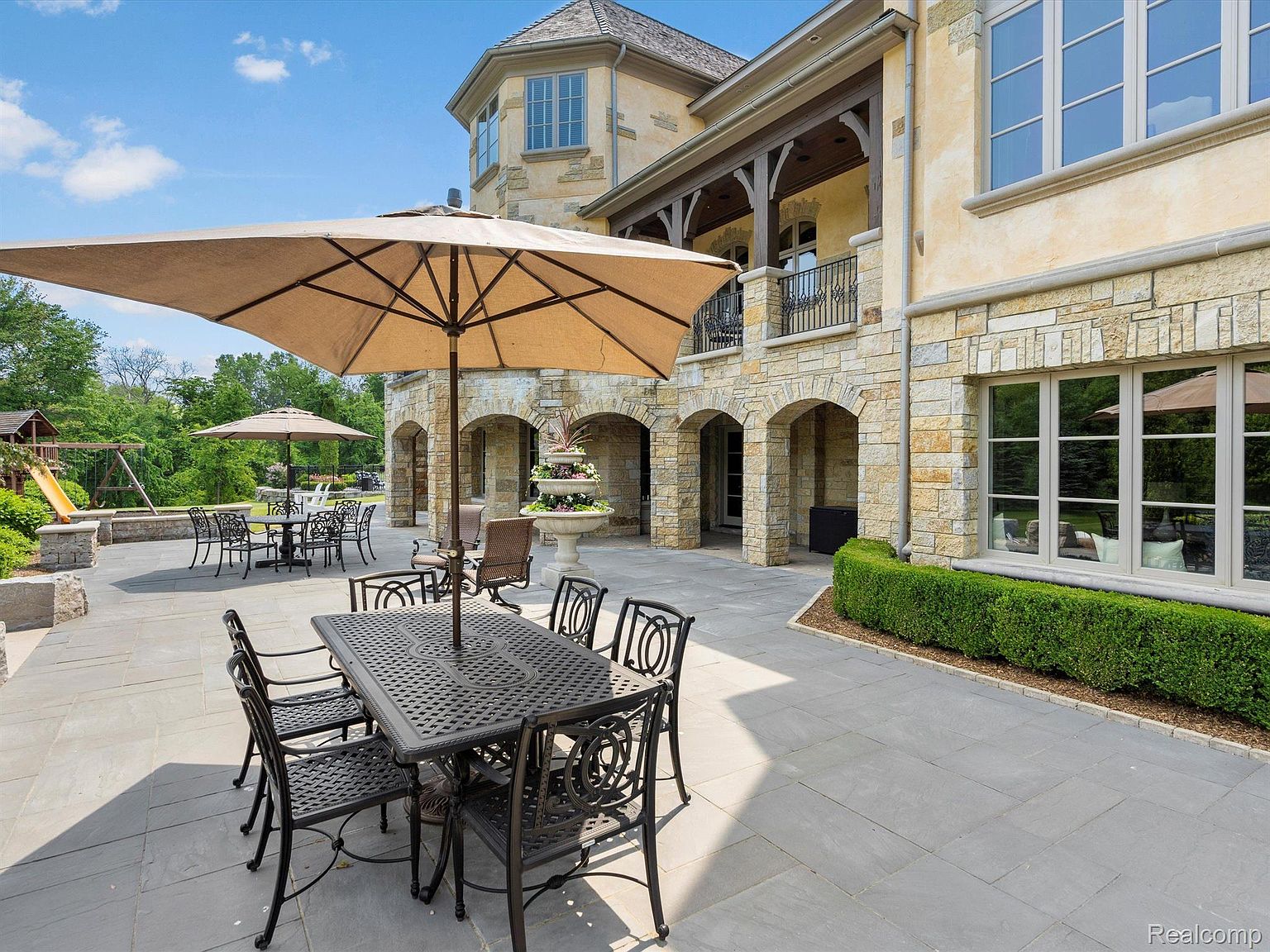 This is a rear exterior view showcasing a spacious stone patio with multiple seating areas, including a large table with chairs under a wide umbrella. The patio is adjacent to a stone-clad building with arched openings and a balcony, suggesting a luxurious outdoor living space. A well-maintained hedge borders the patio, adding to the property's appeal.