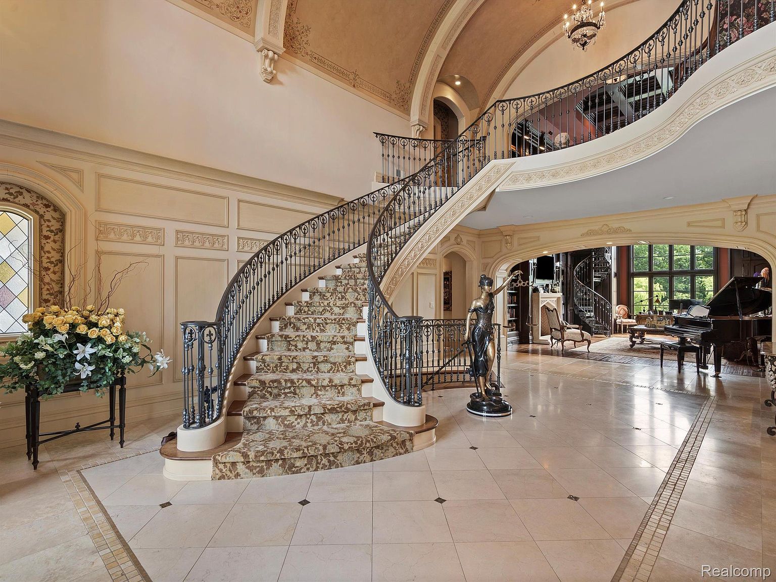 This grand foyer showcases a sweeping staircase with ornate iron railings and patterned carpeting. The space is illuminated by natural light filtering through a stained-glass window, complemented by elegant wall paneling and a polished marble floor. A bronze statue adds a touch of classical artistry, enhancing the luxurious ambiance of the home's entrance.