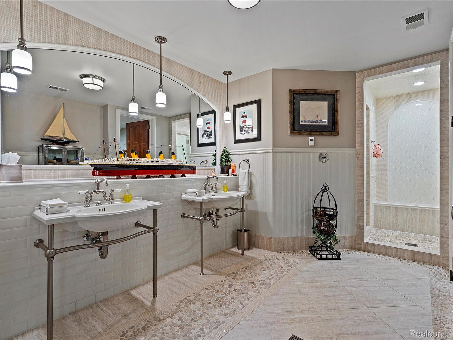 This is a well-appointed primary bathroom featuring dual vanity sinks with chrome fixtures and a unique tile design that transitions from large format tiles to pebble accents. A large mirror reflects the space, enhancing the sense of openness, and a walk-in shower is visible in the background. The bathroom exudes a luxurious and sophisticated ambiance.