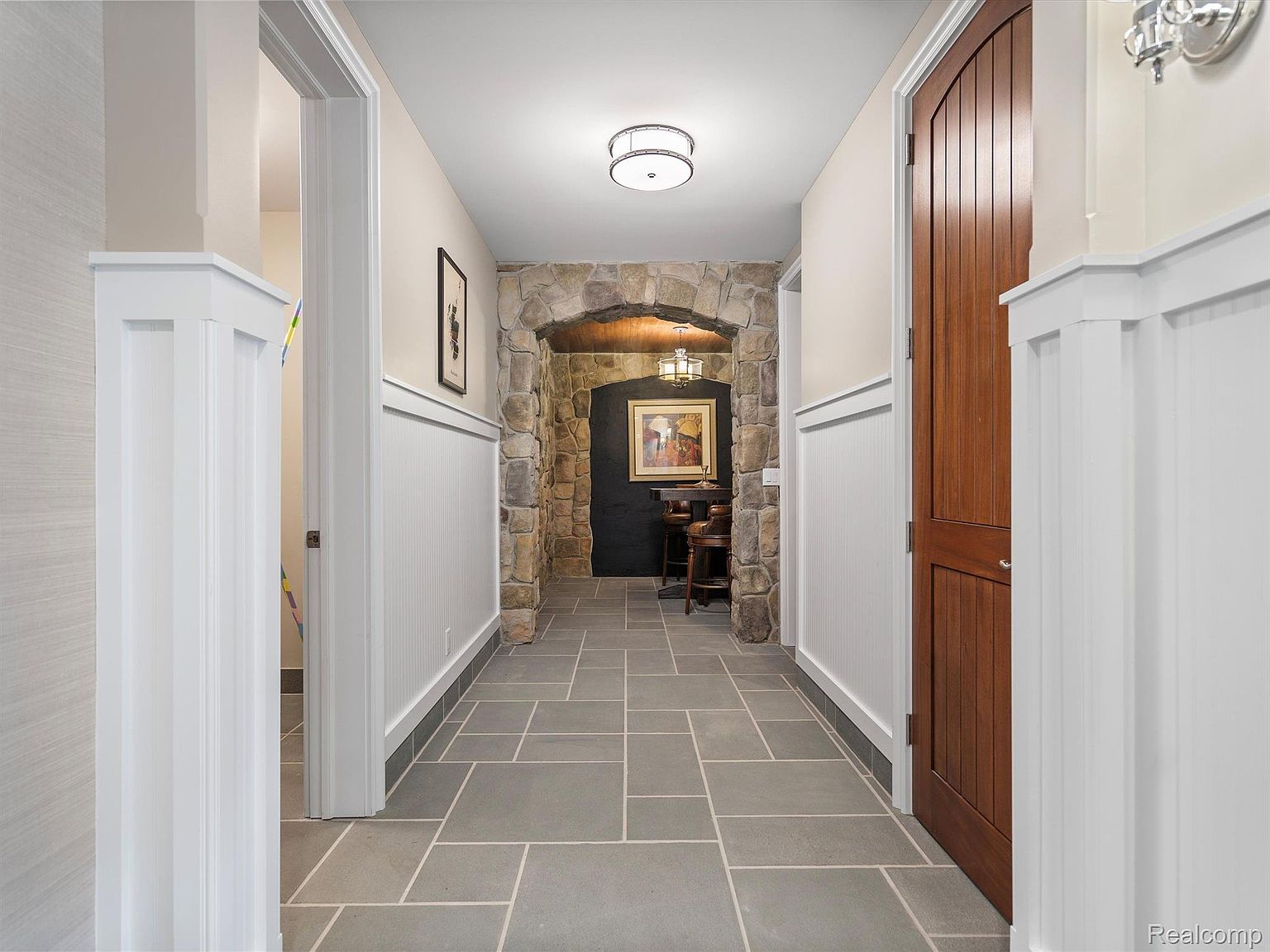 This interior shot showcases a well-lit hallway with a blend of traditional and rustic design elements. The hallway features gray tiled flooring, white wainscoting, and a stone archway leading to a cozy nook with a painting and seating. A wooden door adds warmth, while the overall impression is one of refined comfort and character.