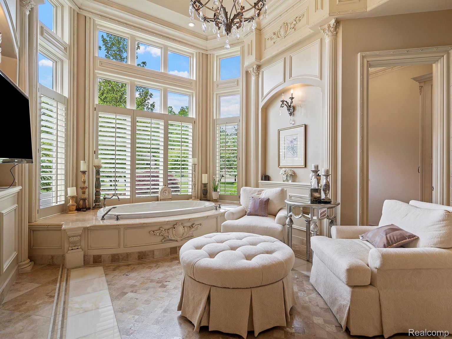 This is a luxurious primary bathroom featuring a large soaking tub situated beneath expansive windows with plantation shutters. The room is elegantly decorated with ornate molding, a crystal chandelier, and comfortable seating, creating a spa-like atmosphere. The flooring is a patterned tile, and the color palette is soft and neutral, enhancing the sense of relaxation and opulence.