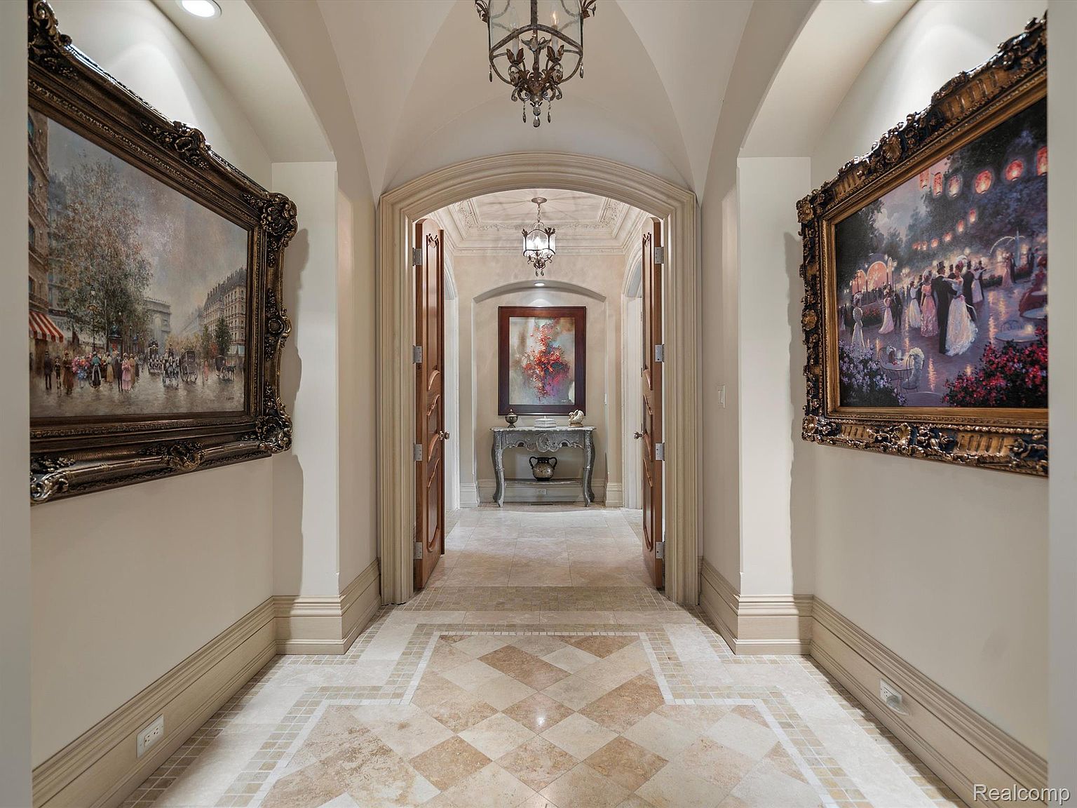 This is an interior shot of a luxurious hallway featuring a barrel-vaulted ceiling, ornate chandeliers, and large framed paintings on either side. The flooring is a decorative tile pattern, and an arched doorway leads to another room with a console table and artwork. The overall impression is one of elegance and sophistication.