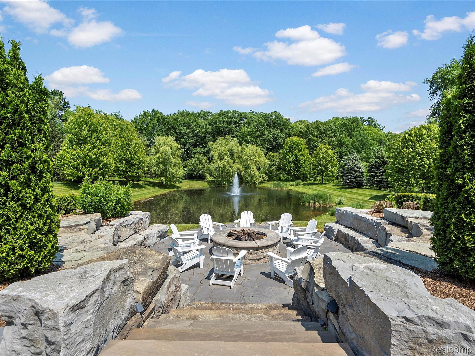 This exterior shot showcases a beautifully landscaped yard featuring a serene pond with a fountain, surrounded by lush greenery and mature trees. A stone patio with a fire pit and white Adirondack chairs creates an inviting outdoor living space. The scene exudes tranquility and highlights the property's potential for relaxation and entertainment.