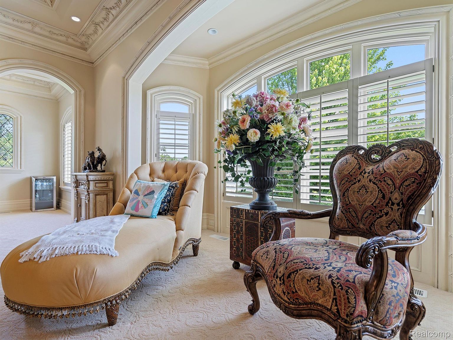 This interior shot showcases a luxurious living room with a chaise lounge, an ornate chair, and a large floral arrangement. The room features arched windows with shutters, detailed ceiling trim, and a neutral color palette, creating an elegant and inviting atmosphere. The overall impression is one of sophistication and comfort.