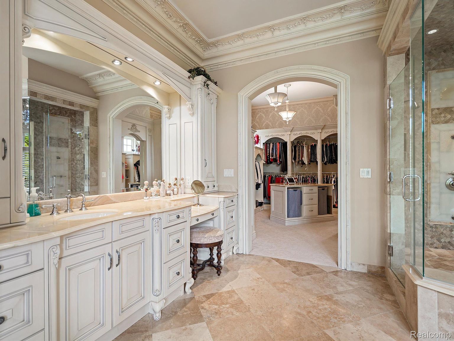 This is a luxurious primary bathroom featuring a vanity with a marble countertop, ornate white cabinetry, and a decorative mirror. The bathroom has a walk-in closet visible through an arched doorway and a glass-enclosed shower. The flooring is tiled, and the overall design exudes elegance and sophistication.