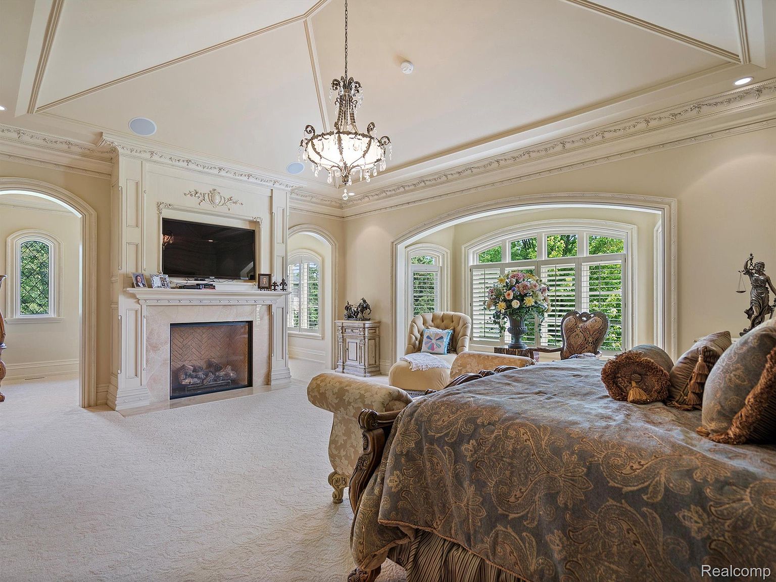 This is a luxurious primary bedroom featuring a large bed with ornate bedding, a fireplace with a TV above, and a seating area by a large arched window. The room is decorated in a classic style with detailed moldings, a chandelier, and neutral colors, creating an elegant and comfortable atmosphere. The perspective is from the foot of the bed, showcasing the room's depth and features.