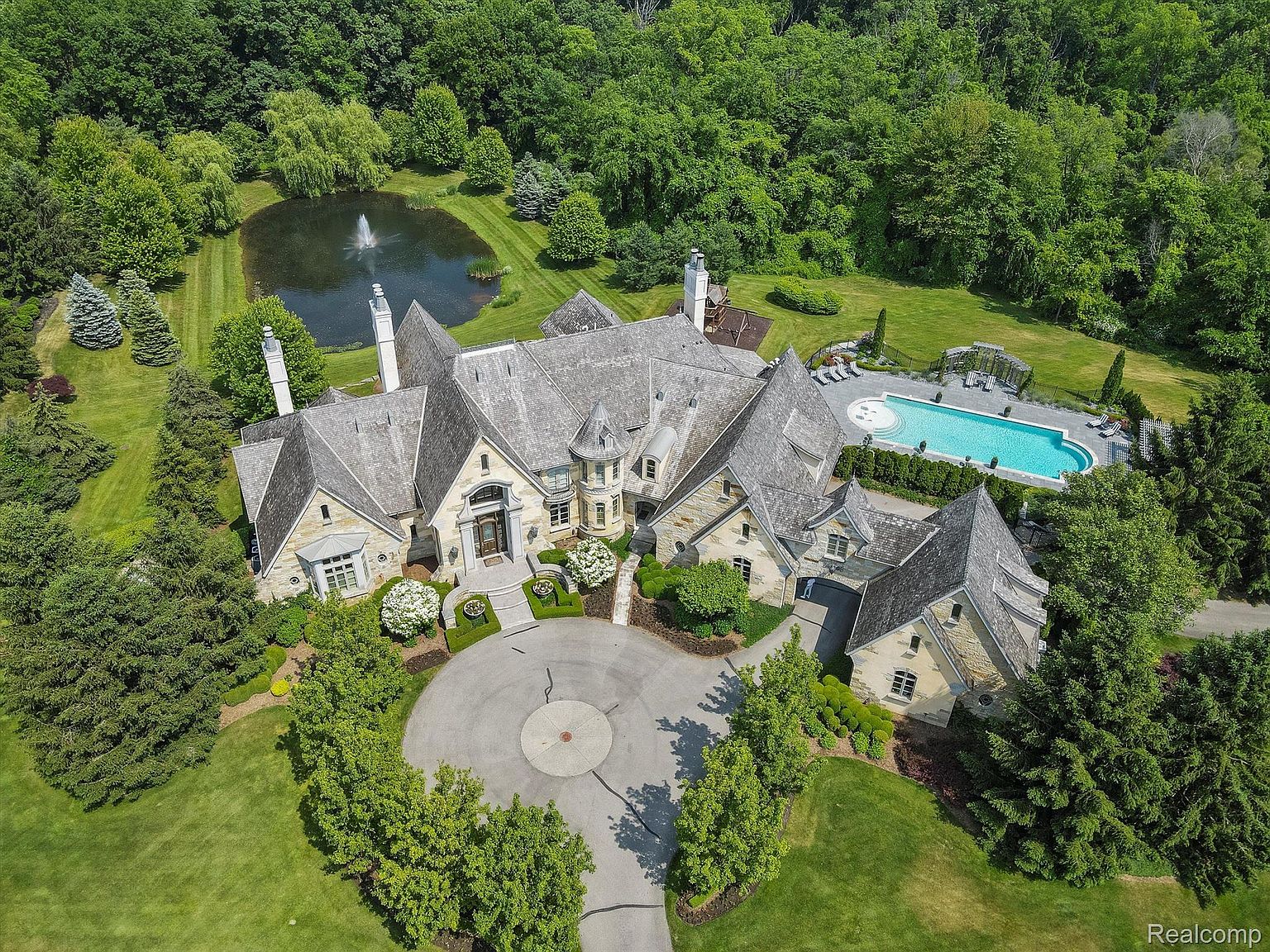 This aerial shot showcases a grand estate with a sprawling layout, featuring a large stone house with multiple gabled roofs and chimneys. A circular driveway leads to the entrance, surrounded by manicured lawns and mature trees. The property also includes a pond with a fountain and a swimming pool with lounge chairs, all set against a backdrop of lush forest, creating a luxurious and private setting.