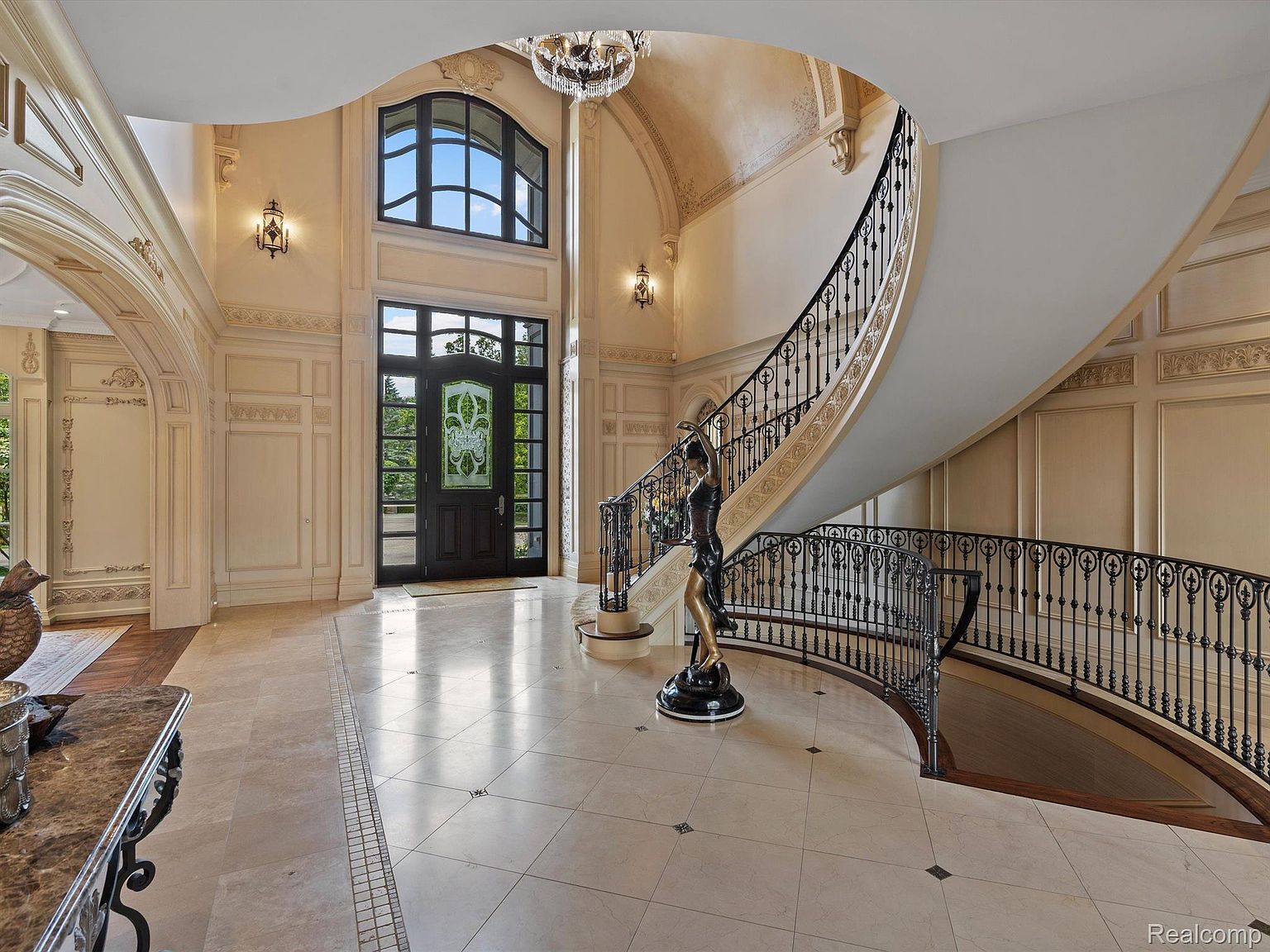 This grand foyer features a sweeping staircase with ornate iron railings, a high ceiling with a decorative chandelier, and elegant wall paneling. The large front door with decorative glass allows natural light to flood the space, highlighting the light-colored tile flooring. A statue adds a touch of sophistication to the entrance.
